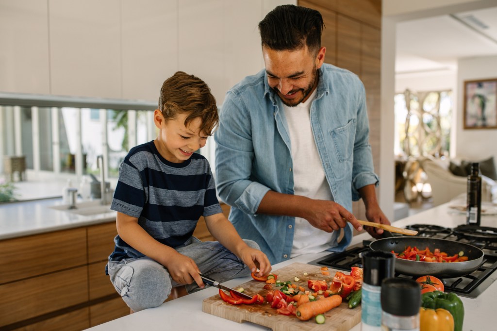 Father and son cooking in the home. Father at the stove while child chops vegetables. Both appear happy.