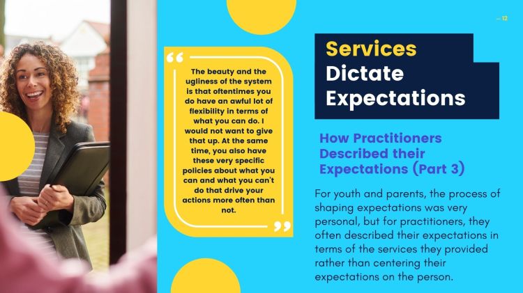 A young woman standing holding a black folder. She is wearing a grey blazer and a striped shirt.