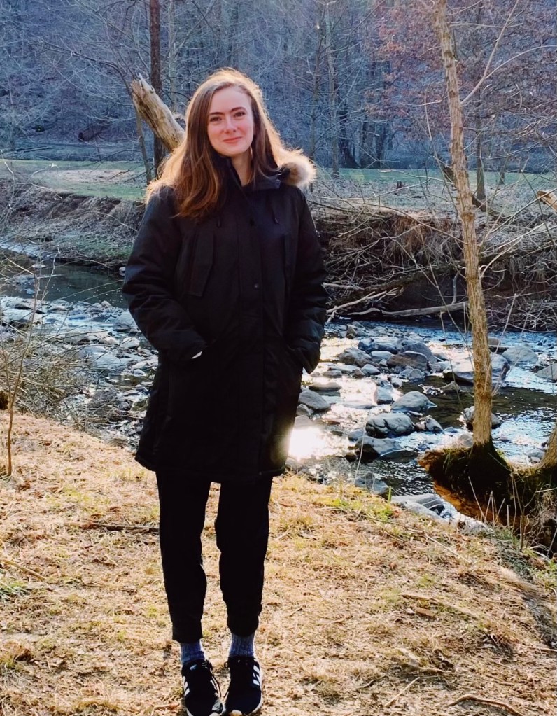 Picture of a young White woman with brown hair wearing a coat standing in front of a stream in winter.