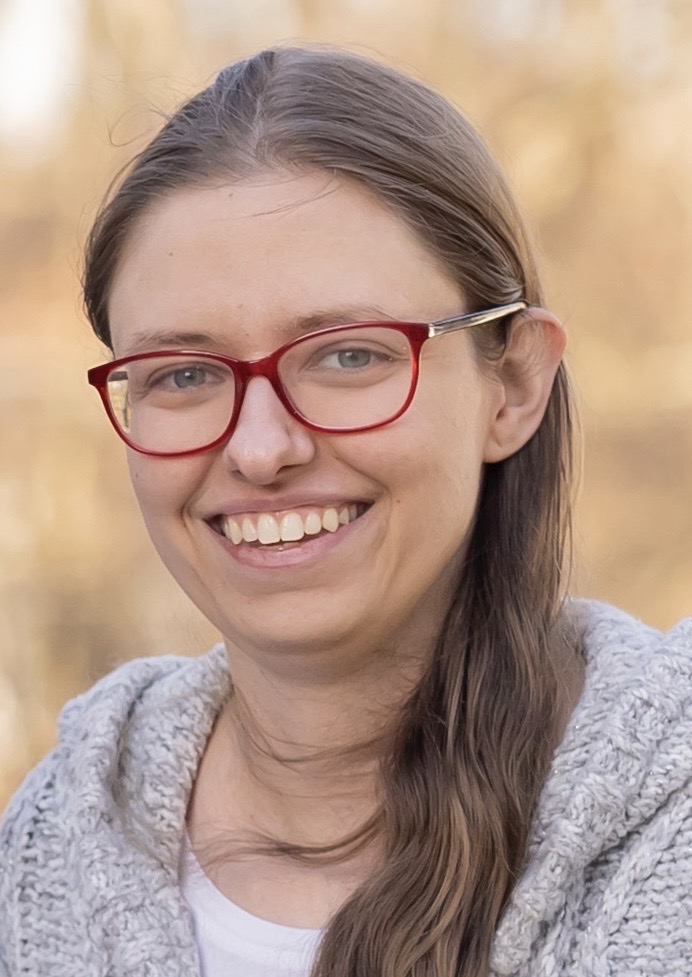 A white woman with blond hair and red glasses smiling. Nature blurred in the background.